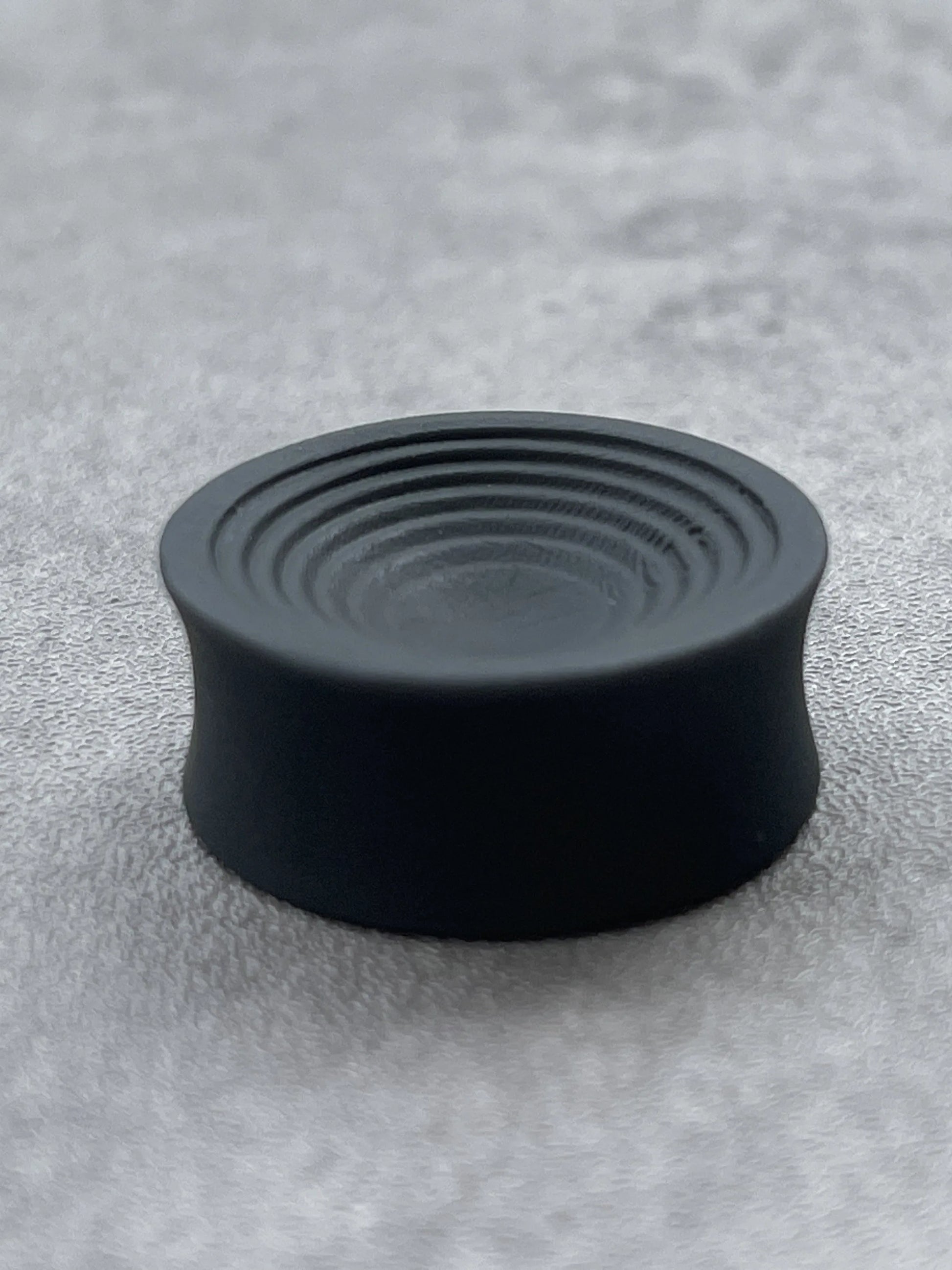 Single black Ripple double flared ear plug on a gray surface